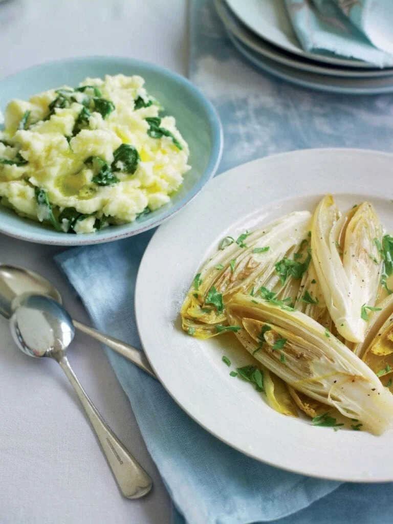 Dusená zelenina na masle na tanieri s vedľa položenými lyžicami a tanierikom so šťouchanými zemiakmi.