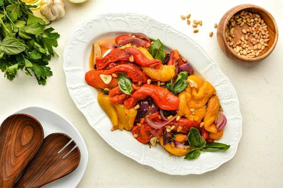 Paprikový šalát s restovanou cibuľou, praženými píniovými orieškami a čerstvými bylinkami servírovaný na oválnom tanieri s vedľa položenými varičkami a miskou semienok.