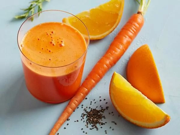 Pomarančovo-mrkvový džús naliaty v pohári, ktorý je položený vedľa čerstvej mrkvy a pomarančov.