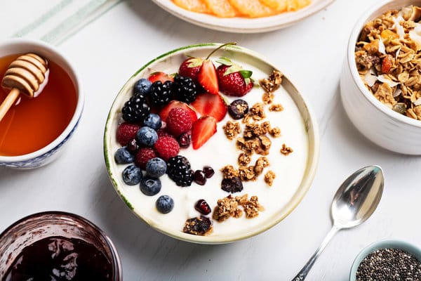 Jogurt vyrobený po domácky servírovaný v miske s müsli a čerstvým ovocím, s vedľa položenou lyžičkou a mištičkami s müsli a sirupmi.