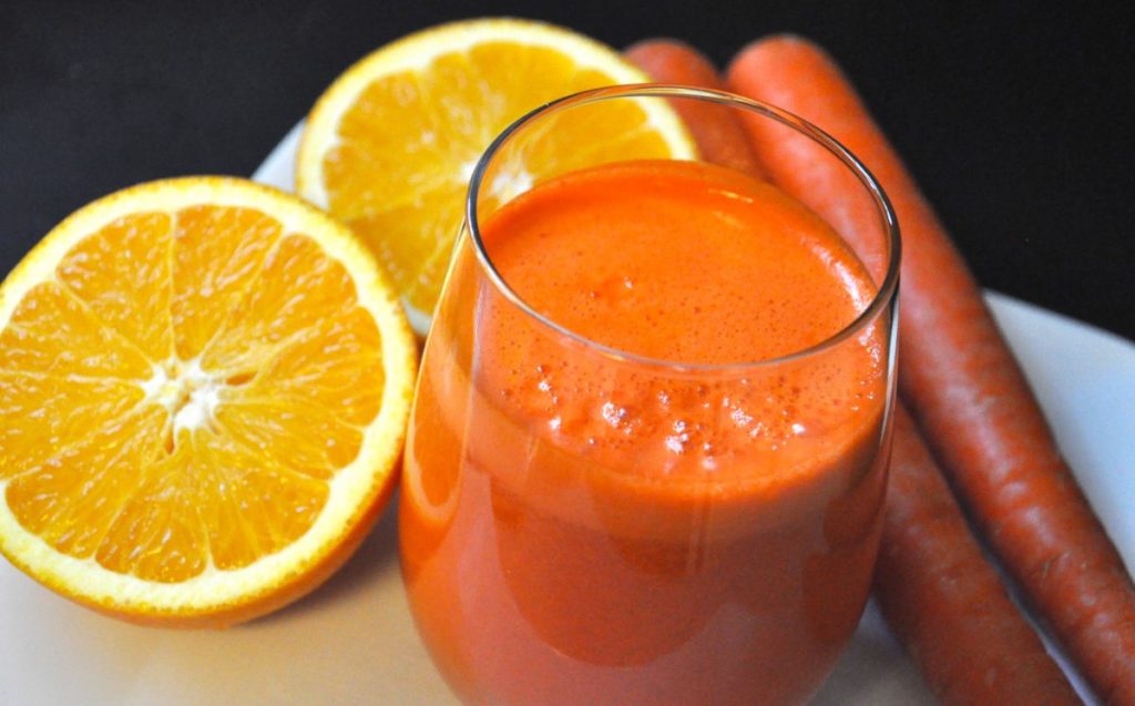 Džús z pomarančov v pohári a vedľa je položený rozpolený pomaranč a dve mrkvy.