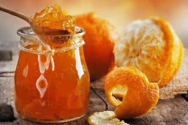 Džem z pomarančov v pohári naberaný lyžicou a vedľa je položený napoly olúpaný pomaranč.