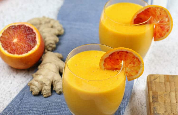 Pomarančovo-zázvorové smoothie v pohároch ozdobené pomarančom s vedľa položeným pomarančom a zázvorom.