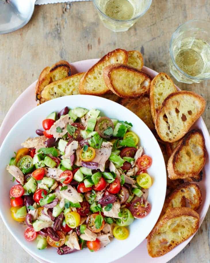 Šalát s tuniakom, uhorkami, paradajkami, olivami a bylinkami servírovaný na tanieri s opečenými plátkami bagety.
