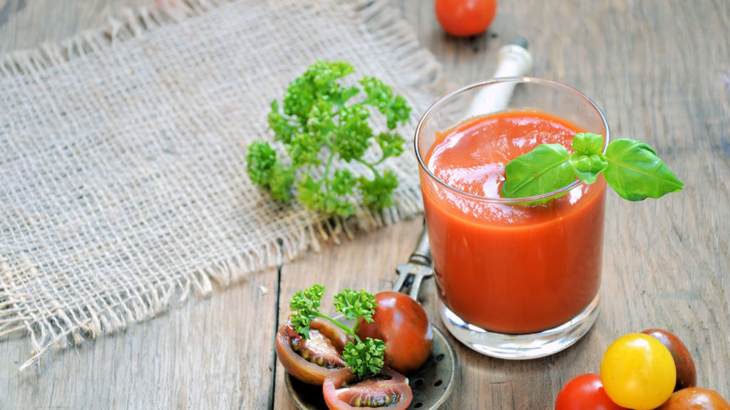 Smoothie z paradajok v pohári, ozdobené čerstvou bazalkou. Vedľa sú položené čerstvé paradajky a bylinky.