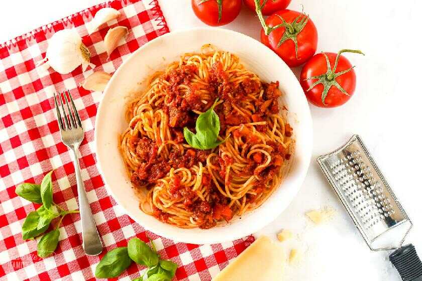 Špagety s mäsom, paradajkami, bazalkou, cibuľou a mrkvou na tanieri s vedľa položeným strúhadlom, paradajkami, cesnakom a bazalkou.