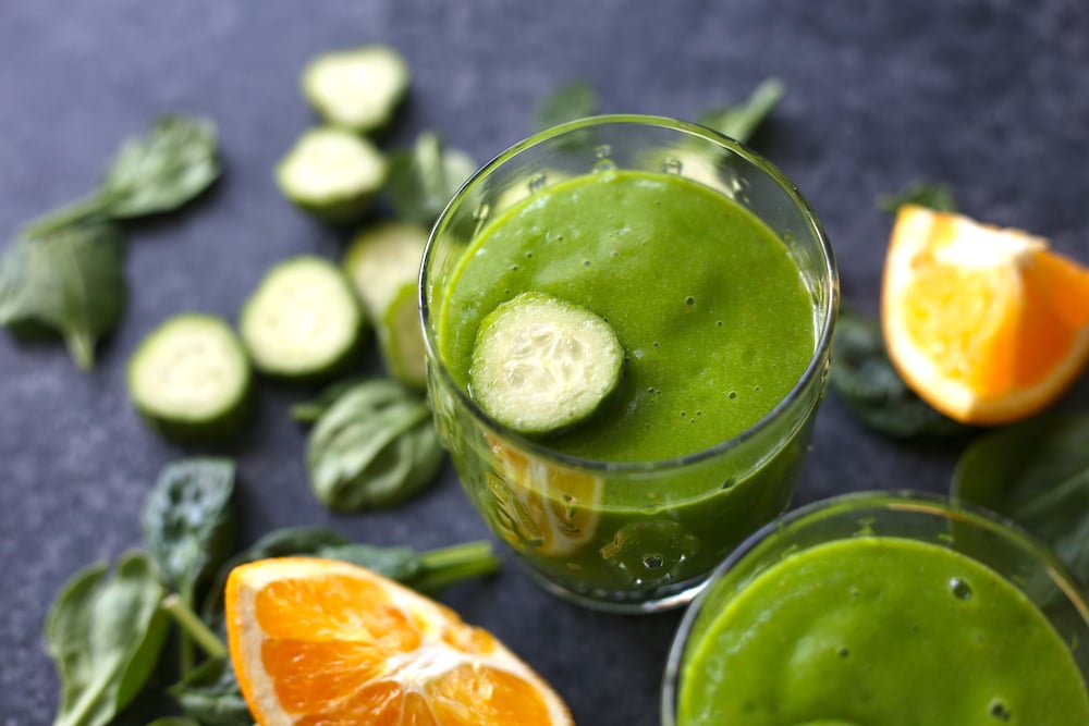 Zelené uhorkové smoothie s čerstvým pomarančom a špenátom v pohároch s plátkom uhorky s vedľa položenými kusmi pomaranča a uhorky.