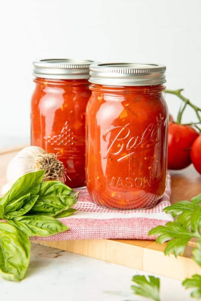 Zakonzervovaná paradajková omáčka v zaváracích pohároch s vedľa položenými listami bazalky, cesnakom a čerstvými paradajkami.
