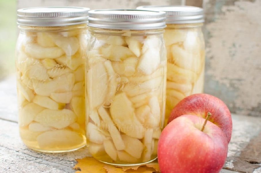 Nakladané jablká v zaváracích pohároch a vedľa položené čerstvé jablká.