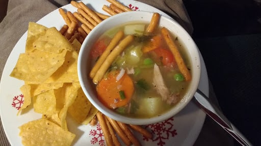 Kuracia polievka s množstvom zeleniny v miske, ktorá je položená na tanieri s tortilla chipsy a slanými tyčinkami.