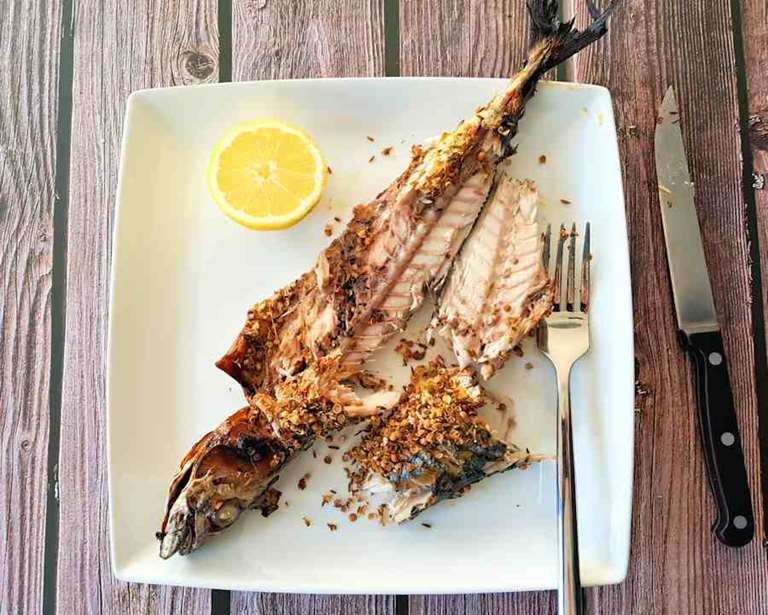 Grilovaná celá makrela s krustičkou z cesnaku, rasce a koriandra na tanieri s vidličkou a plátkom citróna. Vedľa je položený nôž.