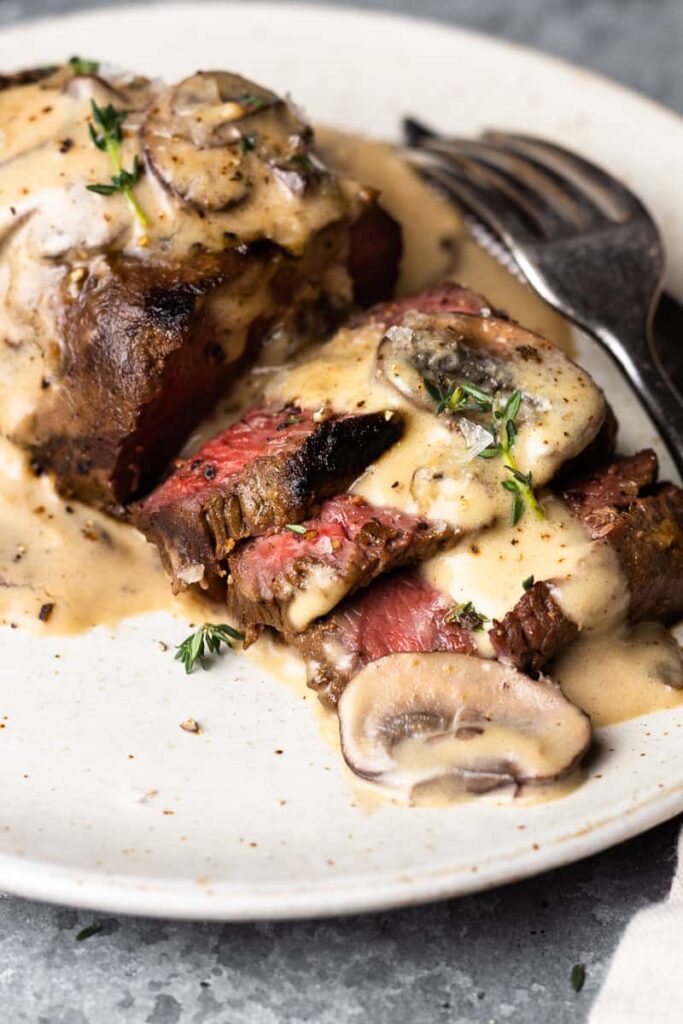 Marinované steaky zo srnčieho mäsa opečené na panvici a podávané s dokonalou krémovou omáčkou zo šampiňónov a bieleho vína na tanieri s príborom.