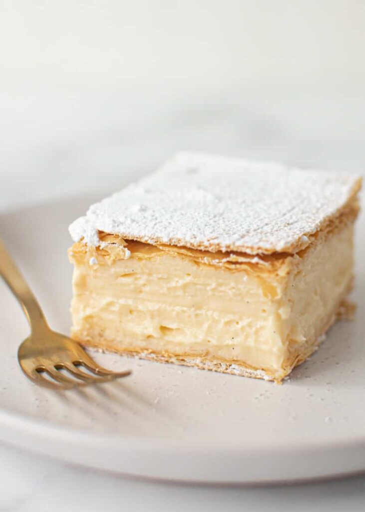 Silná vrstva vanilkového pudingu medzi dvoma chrumkavými plátmi maslového lístkového cesta na tanieriku s vidličkou.
